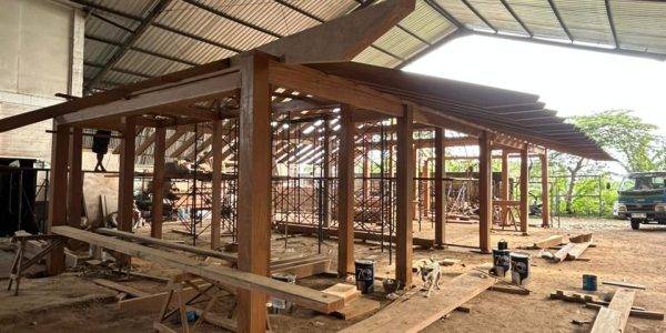“Close perspective of a wooden roof beam system being installed on a Kontiki building mockup, with scaffolding and construction materials on the warehouse floor